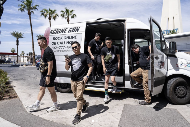 Five men exit a white van with a booking ad on the side, near palm trees.