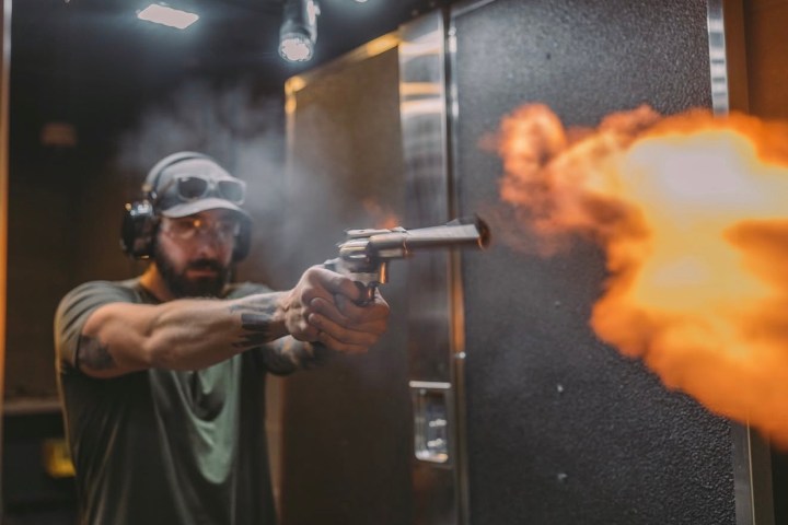 Man firing a handgun at an indoor shooting range with muzzle flash visible.