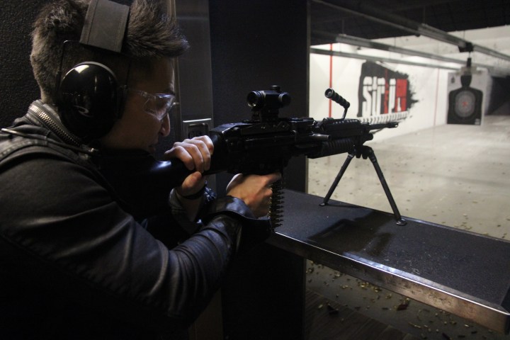 Person aiming a rifle at a shooting range with ear protection and safety glasses.