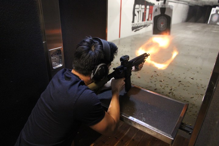 Person firing a rifle at an indoor shooting range, muzzle flash visible.
