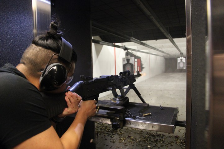 Person firing rifle at indoor shooting range, wearing ear protection.