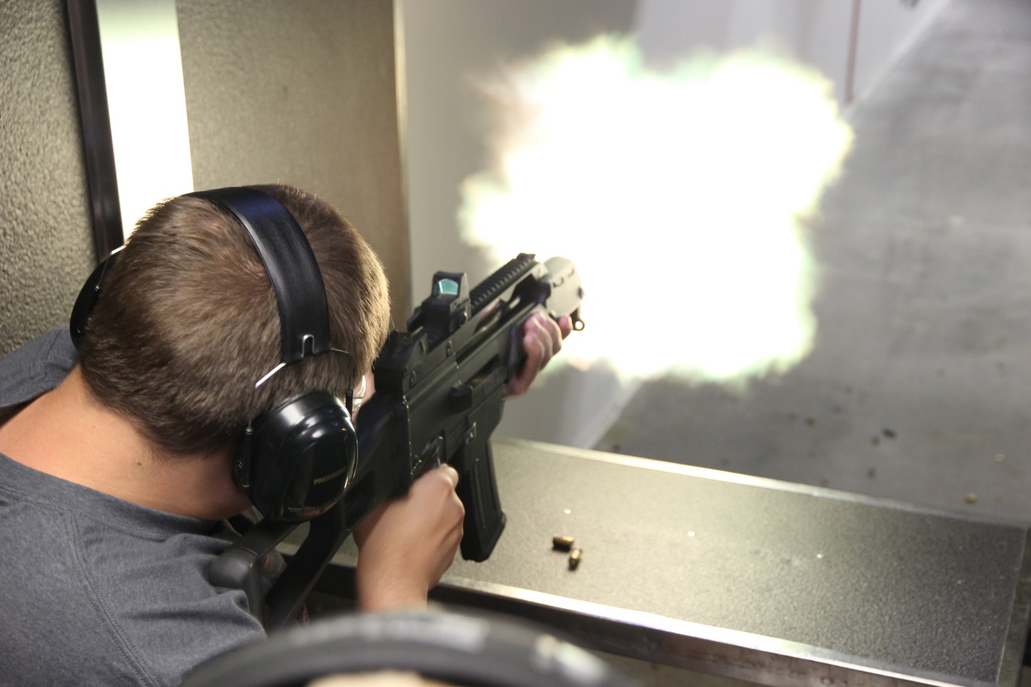 Person firing a rifle at a shooting range, wearing ear protection, with muzzle flash visible.