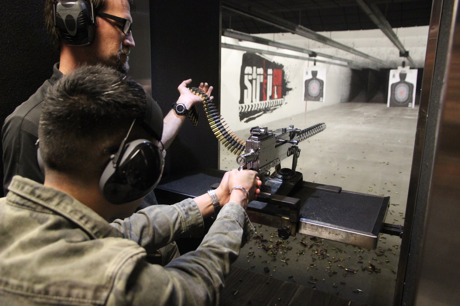 Two people at a shooting range using a mounted machine gun with headphones on.