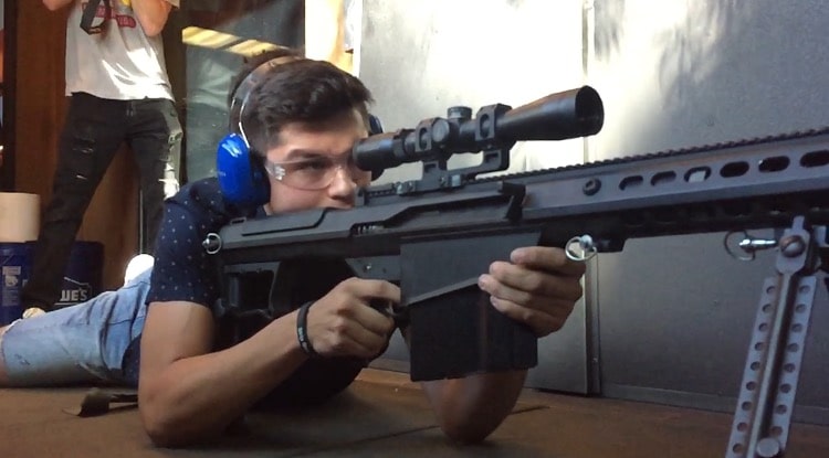 Person lying down aiming a sniper rifle with blue ear protection in an indoor range.