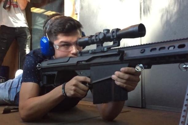 Person lying down aiming a sniper rifle with blue ear protection in an indoor range.
