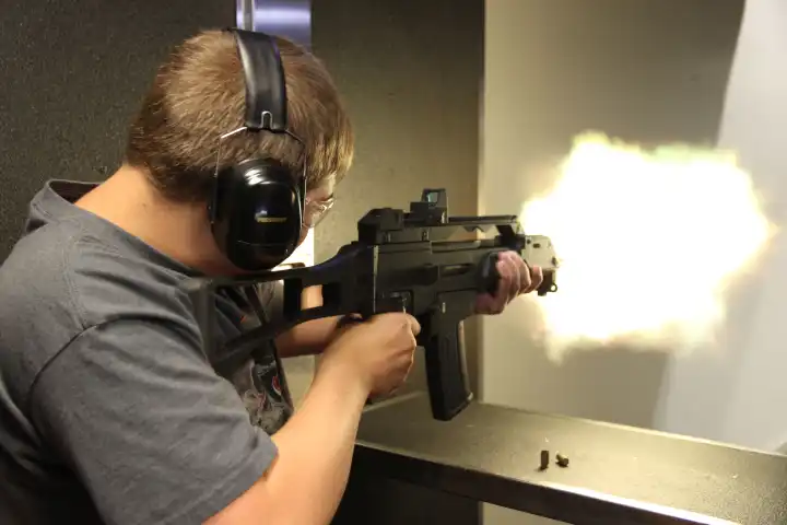 Person firing a gun indoors, wearing ear protection and safety glasses, with muzzle flash visible.