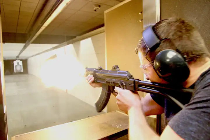 Person firing a rifle at an indoor shooting range, wearing ear protection and safety glasses.
