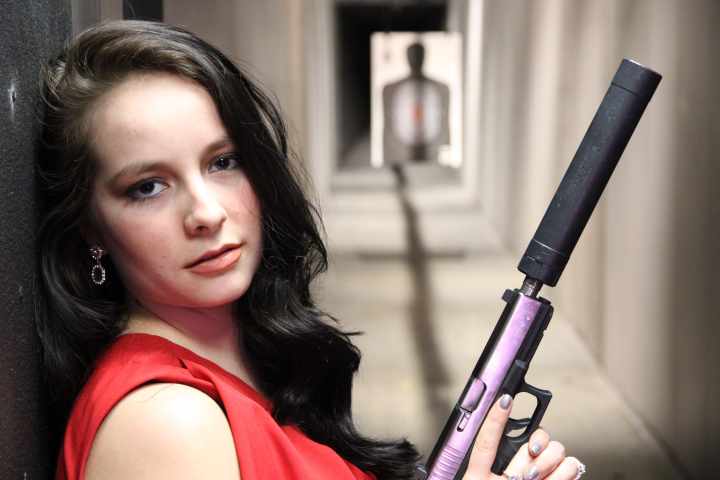 Woman in red dress holding a silenced pistol at a shooting range, target visible in background.