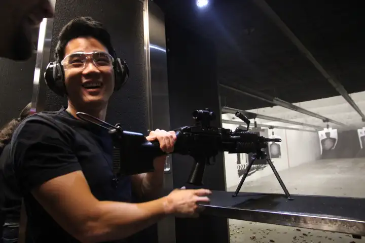 Person smiling at indoor shooting range with ear protection and rifle on stand.